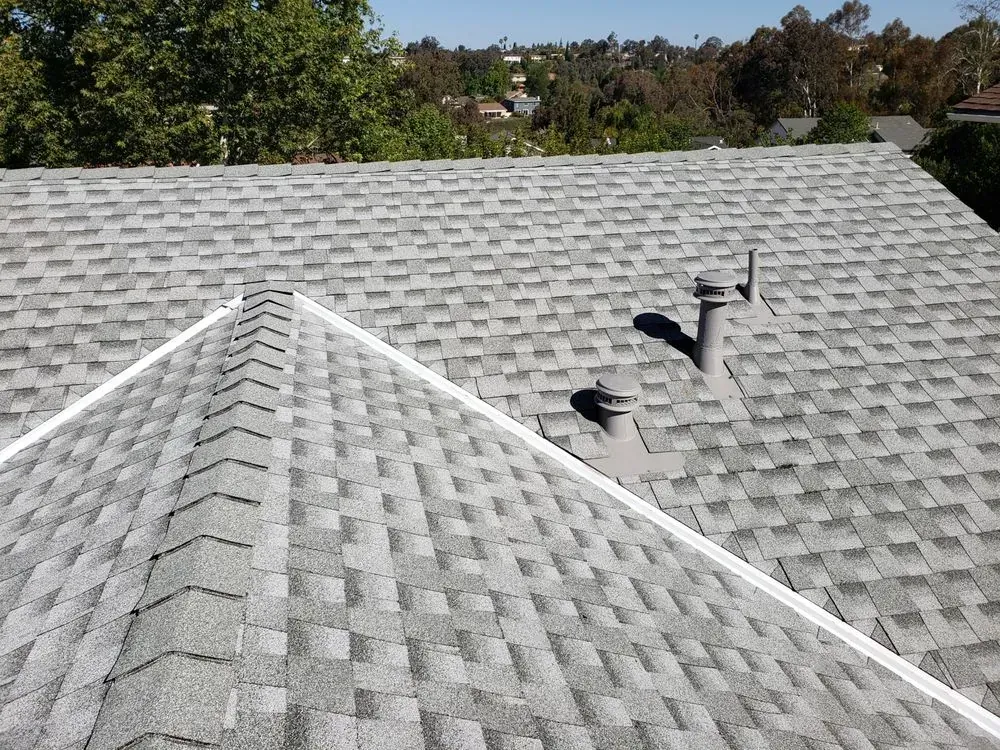 A high-angle view of a grey shingled roof featuring a ridge line, two vent pipes, and surrounding trees in the background.