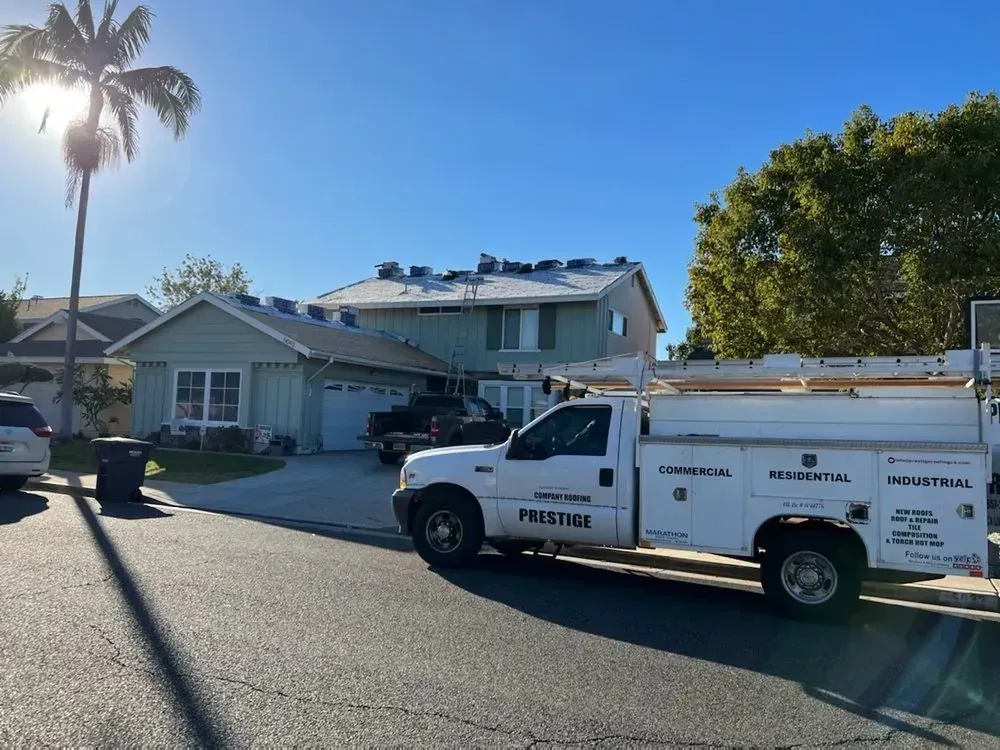 A white service truck is parked on a street in front of a light green house with a residential roof under repair.