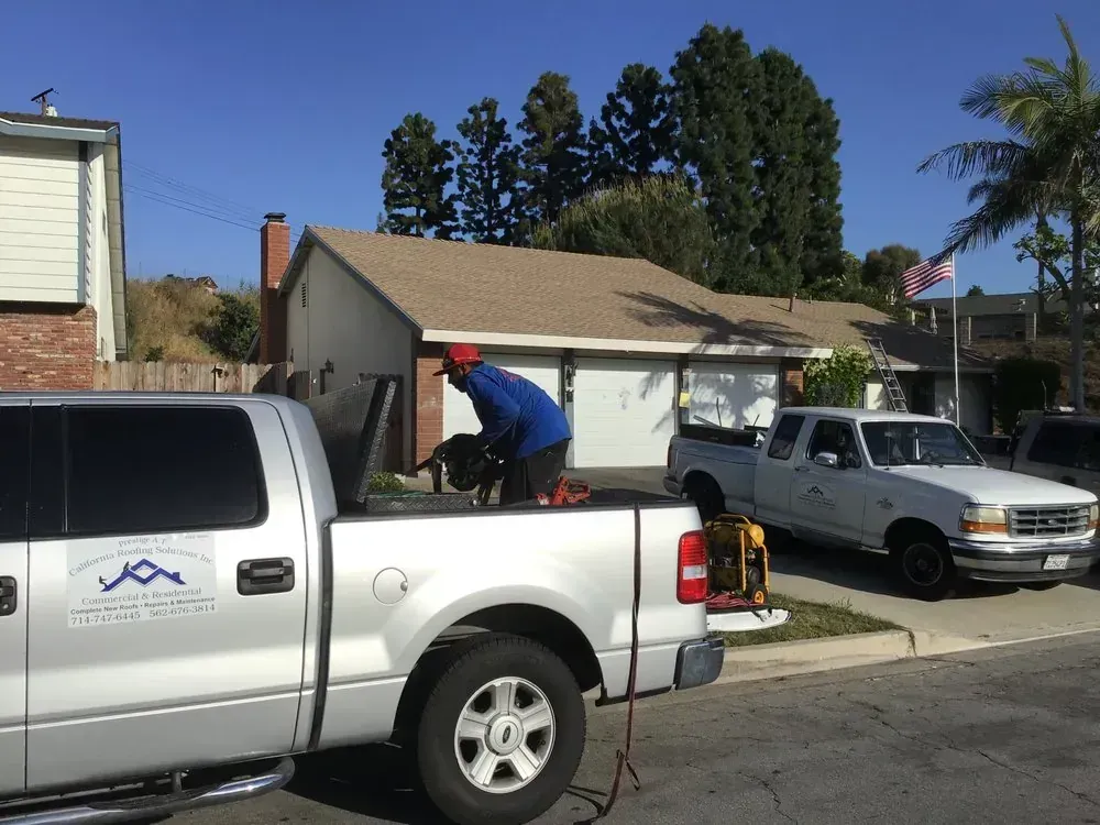 A person in a blue shirt and red cap works near the back of a silver pickup truck parked on a residential street.