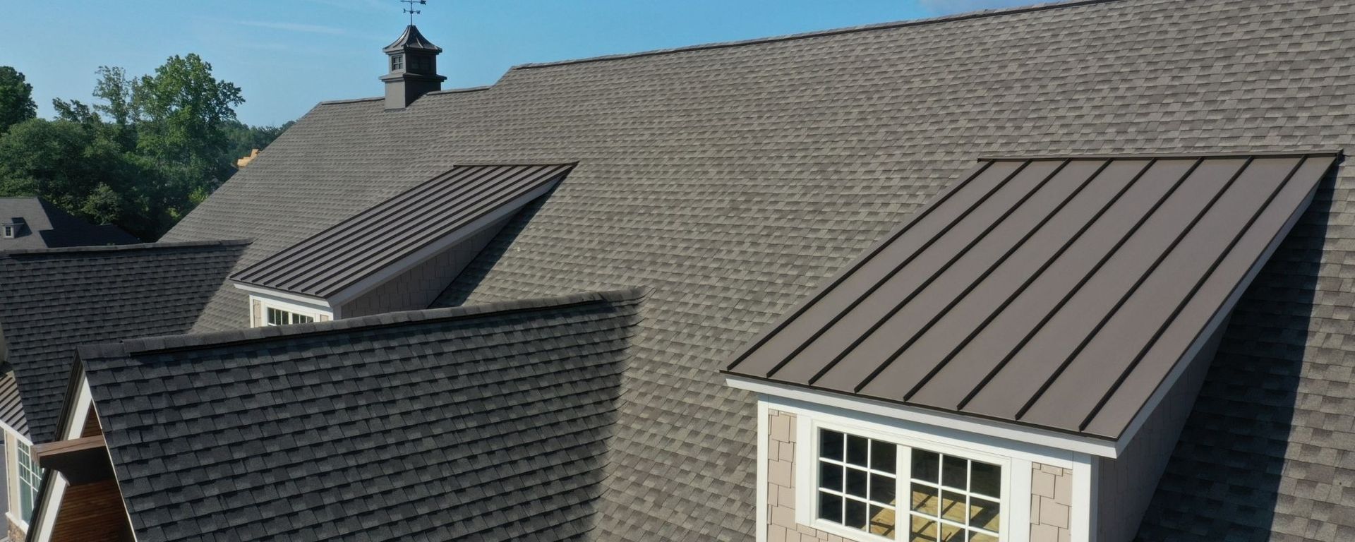 A sloped shingle roof with two brown metal-topped dormers and a small weather vane on a sunny day.