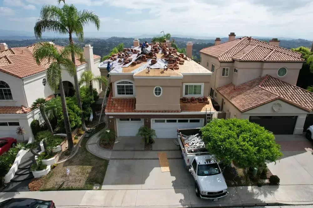 An aerial view of a tan two-story house undergoing a roof replacement with workers visible on the roof.