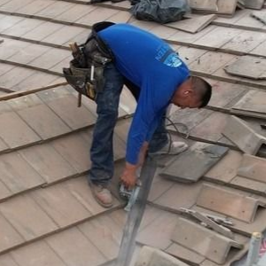 A worker in a blue shirt uses a handheld power saw to cut a tile on a residential roof.