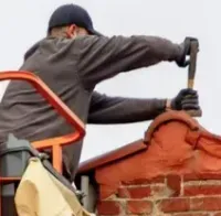 A person in a cap and dark clothing using a hammer to repair a red brick rooftop from an orange aerial lift.