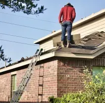 A person in a red long-sleeved shirt and jeans stands on a sloped residential roof near an extension ladder.