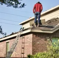 A person in a red long-sleeved shirt and jeans stands on a sloped residential roof near an extension ladder.