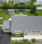 An aerial view of a single-story suburban house with a gray shingle roof, a driveway, a backyard, and a small shed.