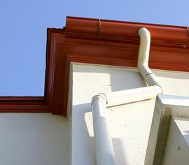 A white downspout attached to the corner of a building with rust-colored gutters under a clear blue sky.