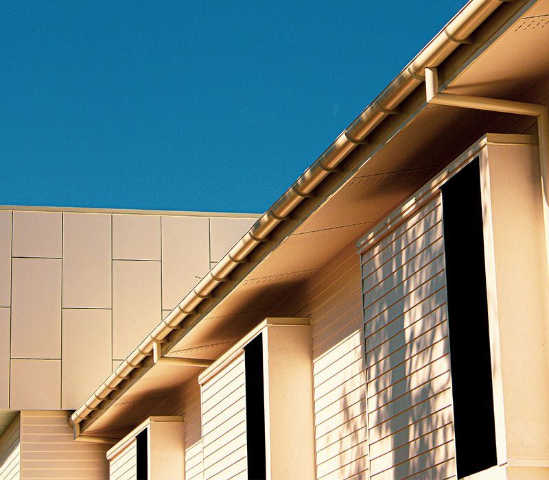 Low-angle view of a modern building exterior with cream-colored siding, a metal gutter, and a clear blue sky.