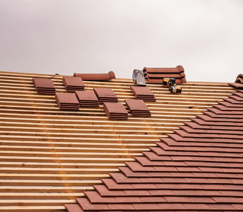 Roof under construction showing rows of wooden battens, stacks of red clay roof tiles, and a power tool on the slope.