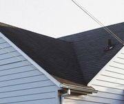A close-up view of the roofline and light-colored siding of a house with black shingles against a bright sky.