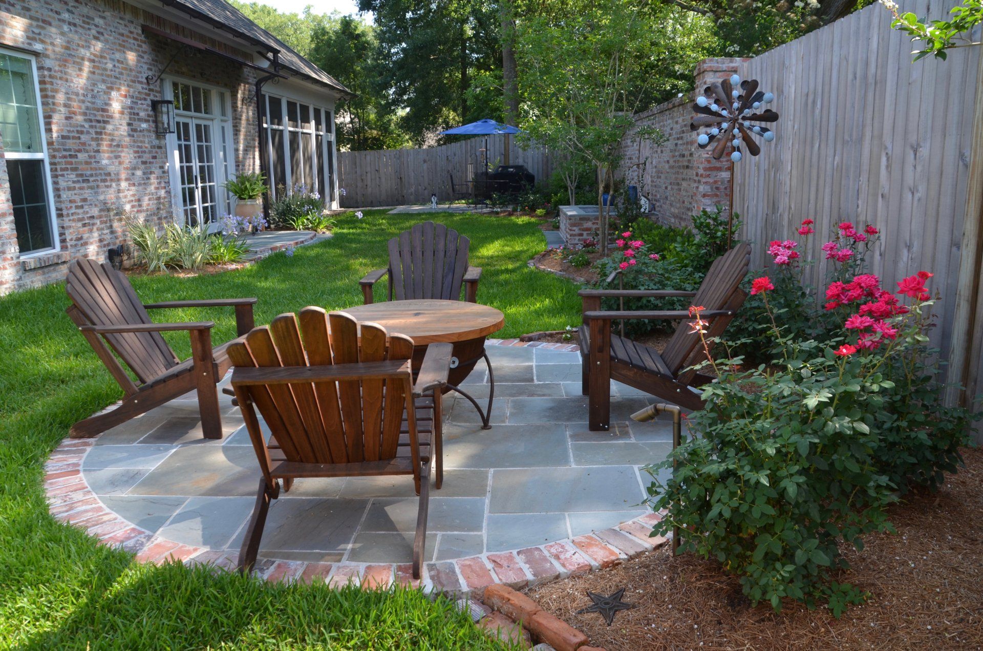 Patio with wooden chairs and table, brick border, and flowers next to a wooden fence.