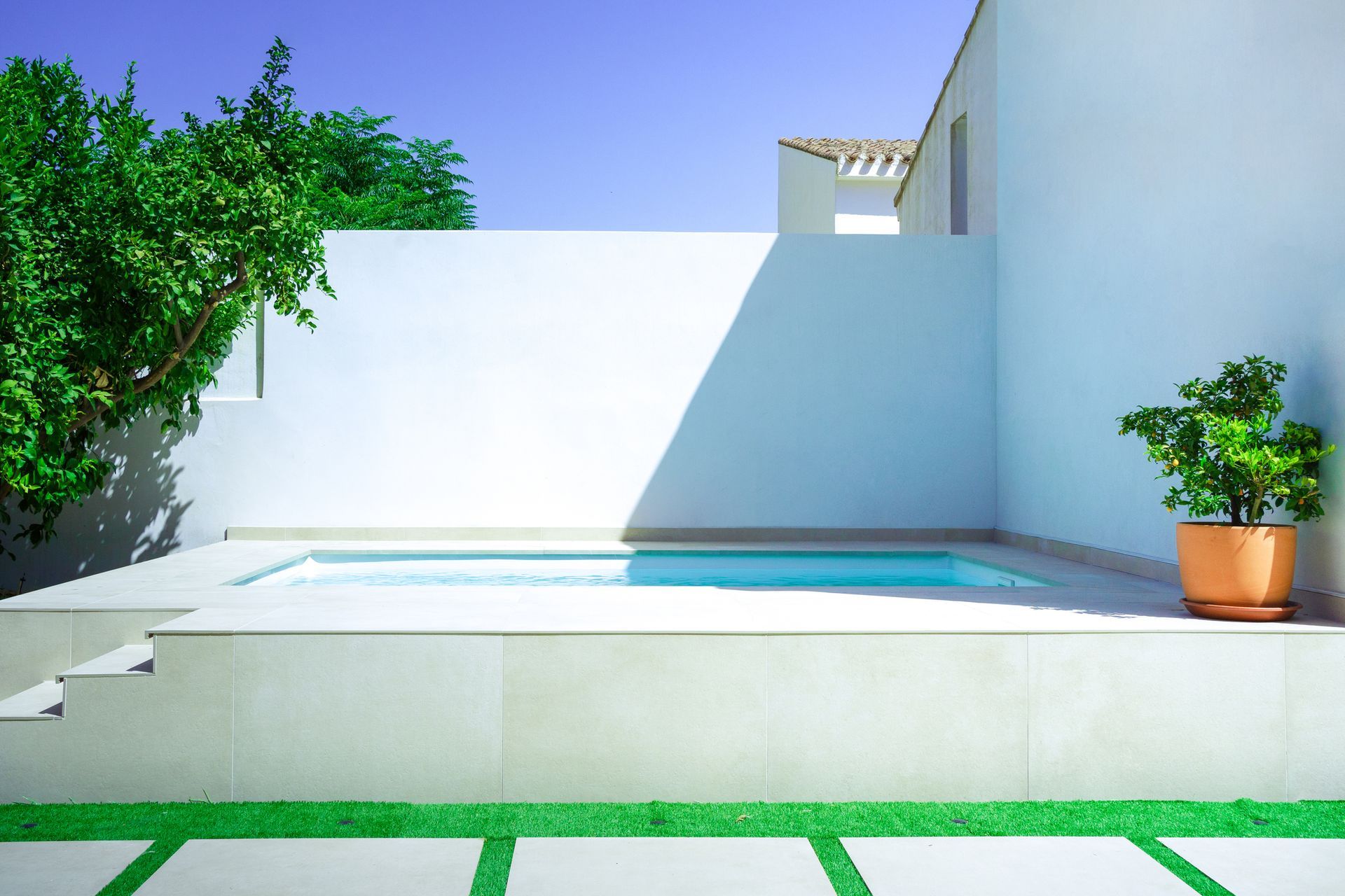 Modern backyard pool with sleek custom concrete patio and white minimalist walls in sunlight.