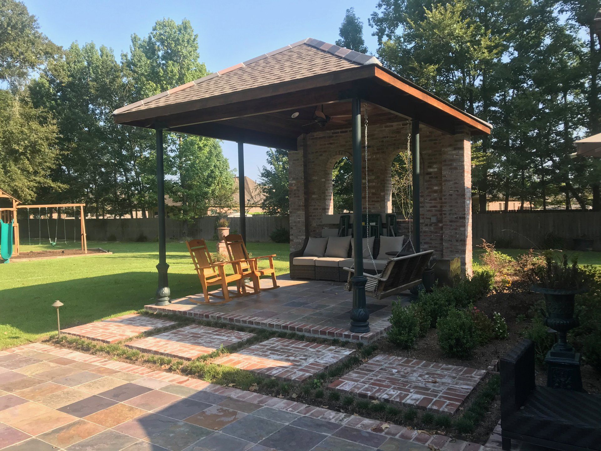 Brick patio with gazebo; seating includes wooden chairs and a swing. Green lawn and trees in background.