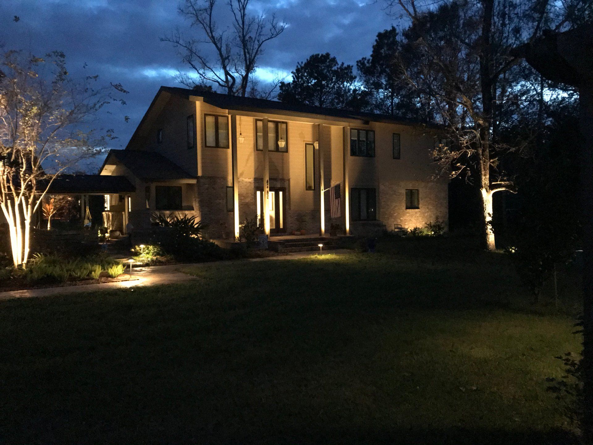 House exterior at night, illuminated by landscape lighting. Beige siding, dark sky, and lawn.