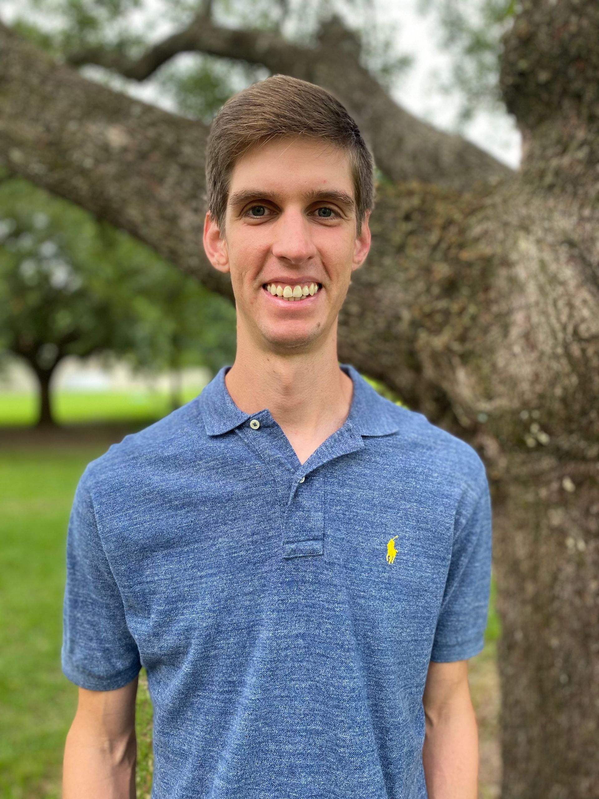 Man smiling in a blue polo shirt, standing in front of a tree outdoors.