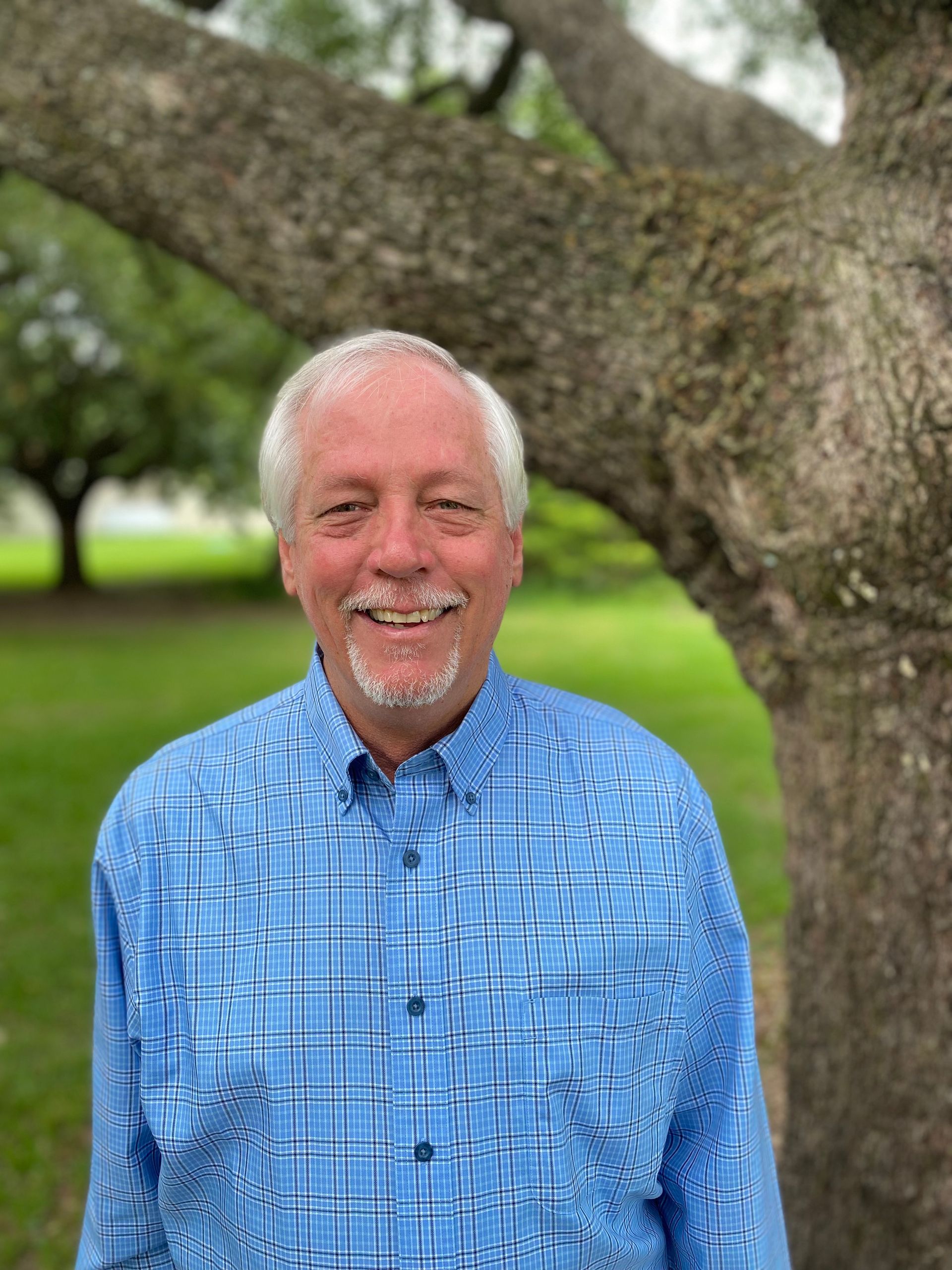 Smiling man in a blue plaid shirt stands outside near a tree.