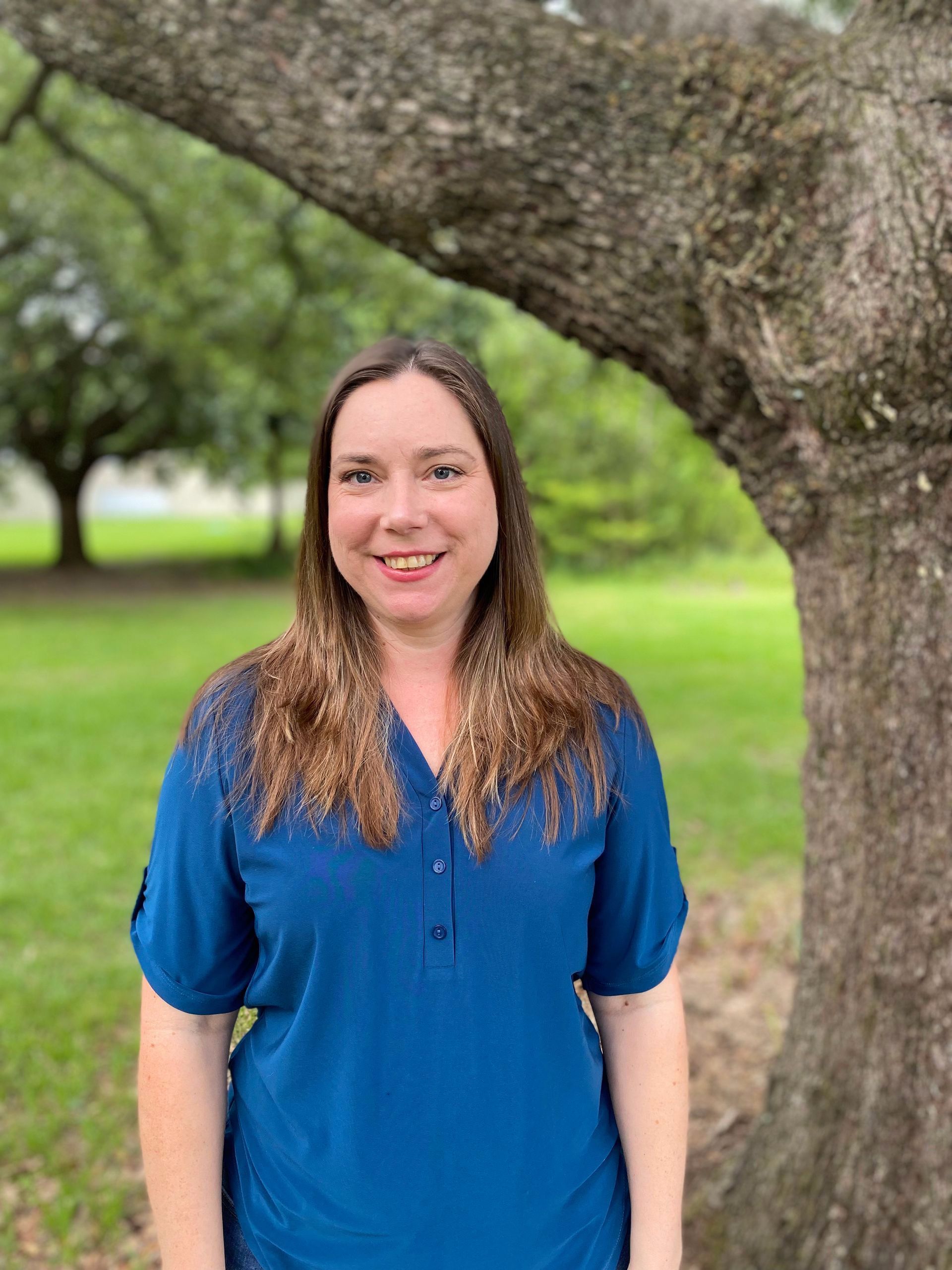 Woman in blue shirt smiles in front of a tree outdoors.