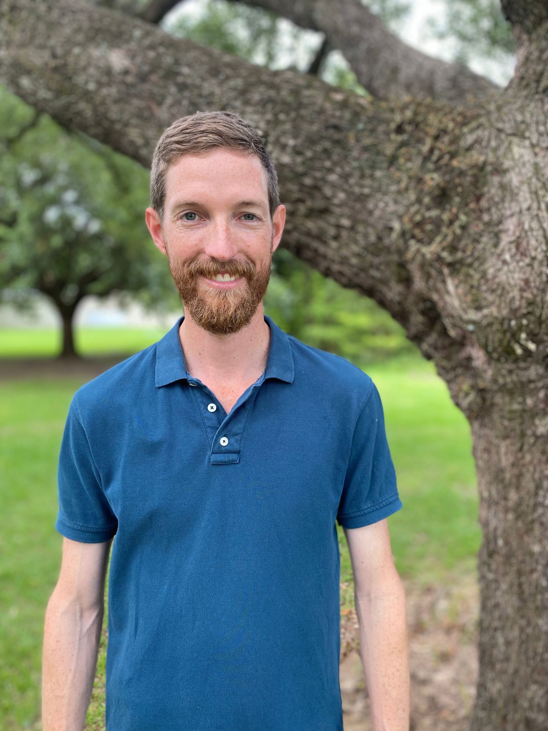 Man with a reddish beard in a blue polo shirt standing near a tree, smiling slightly outdoors.