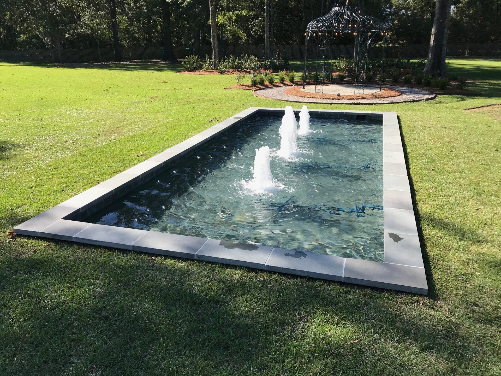 Rectangular fountain with three water jets on a grassy lawn; trees in the background.