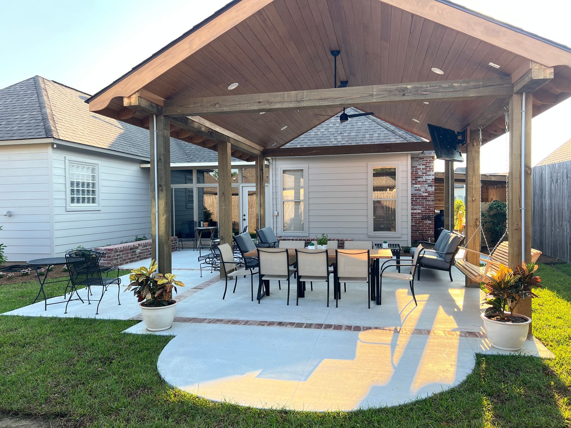 Covered patio with seating, television, and potted plants in a backyard setting.