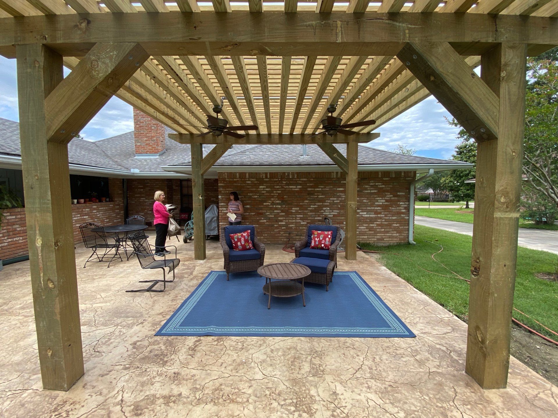 Wooden pergola over a patio with outdoor furniture, two people standing near the house.