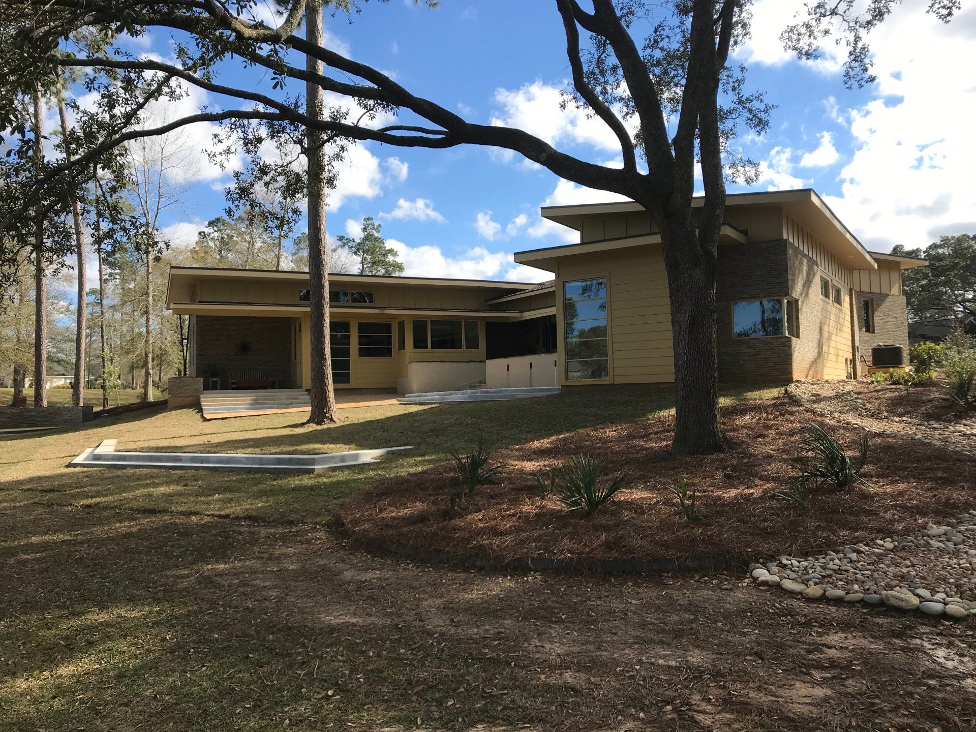 Tan modern house with large windows, surrounded by trees and a lawn under a blue sky.