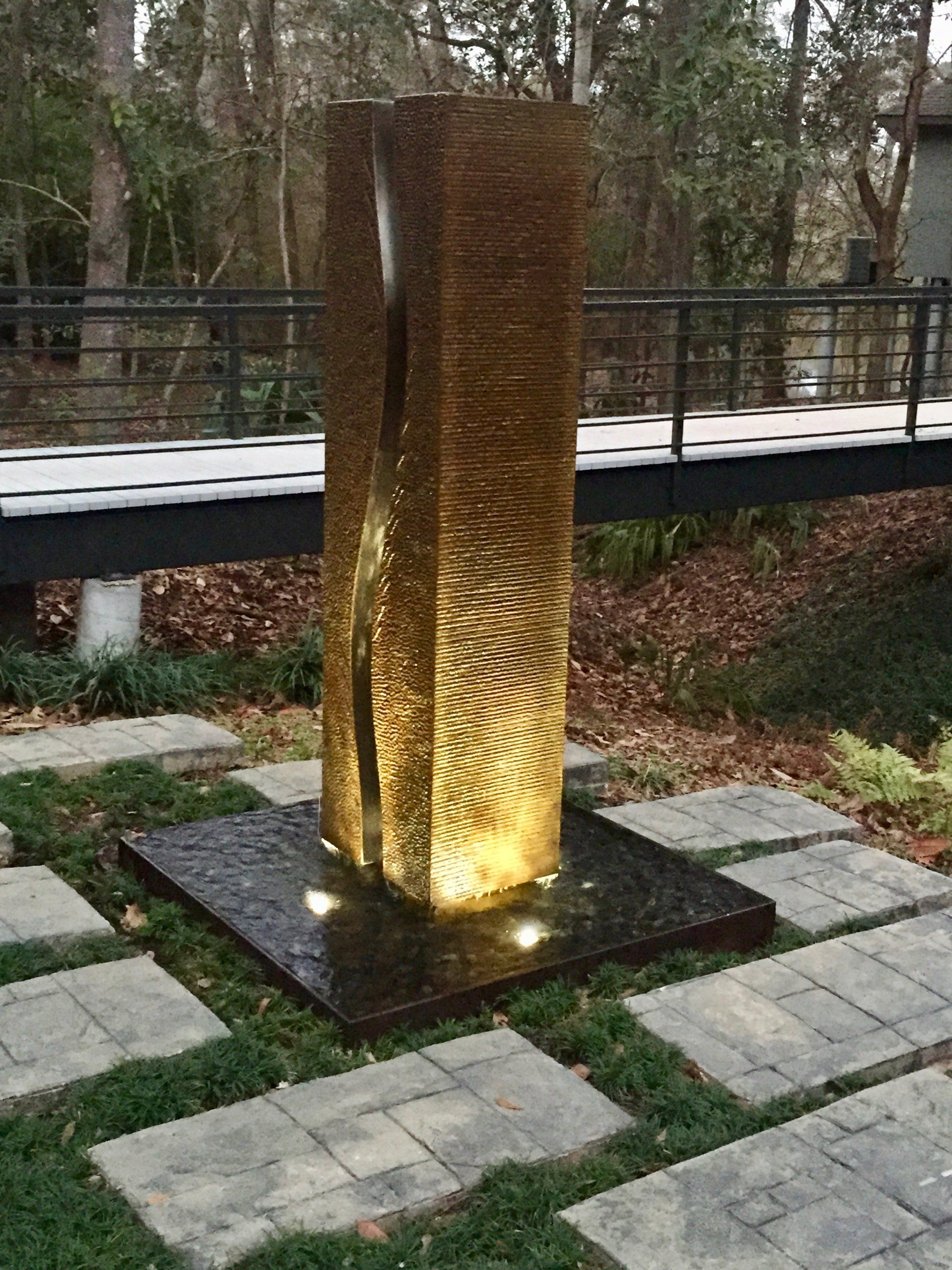 A modern water fountain sculpture in a garden setting. Brown stone with water feature.