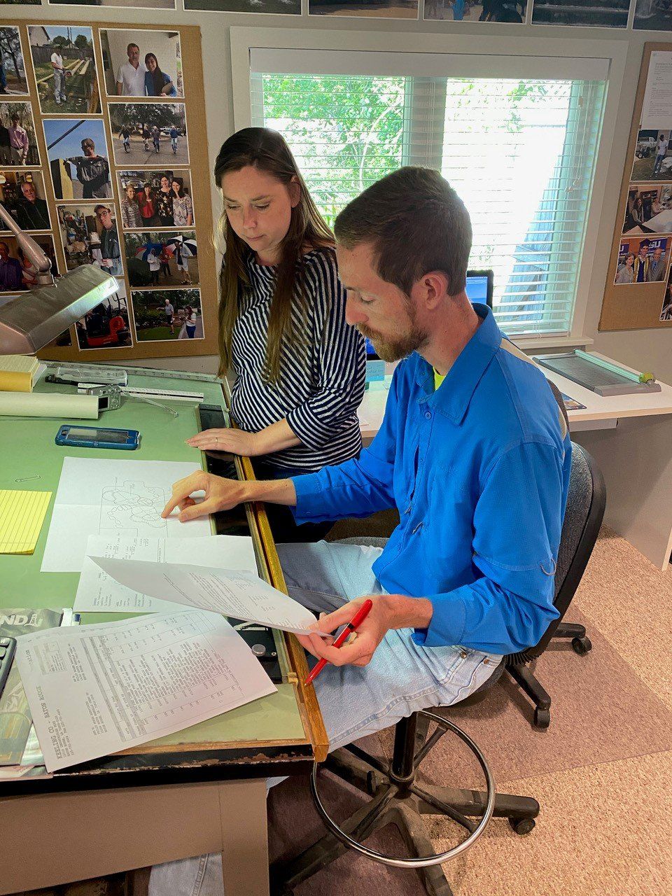 Man in blue shirt and woman in striped shirt reviewing documents at a desk in a workspace with photos.