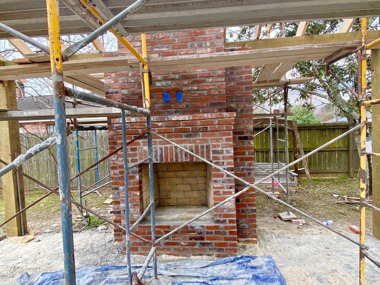 Brick fireplace under construction, surrounded by scaffolding. Outdoors, with a partially visible wooden structure.