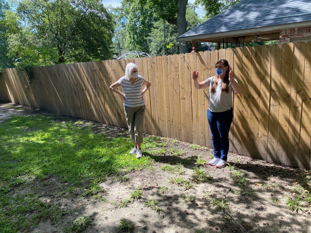 Two people stand near a wooden fence outdoors, one wearing a mask and gesturing, the other with hands on hips.