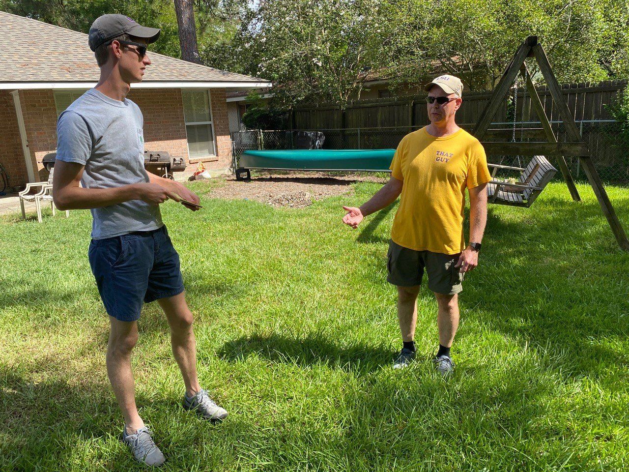 Two men standing in a grassy backyard, conversing. One wears gray shirt, shorts, cap. The other, yellow shirt, shorts, cap. Canoe visible.