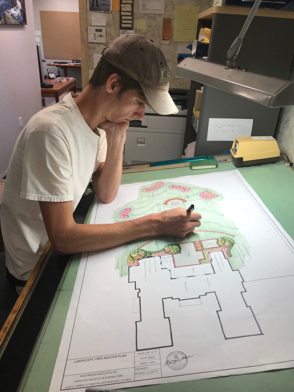 Man in cap drawing a landscape design on a large blueprint table, using a pen to add details and color.