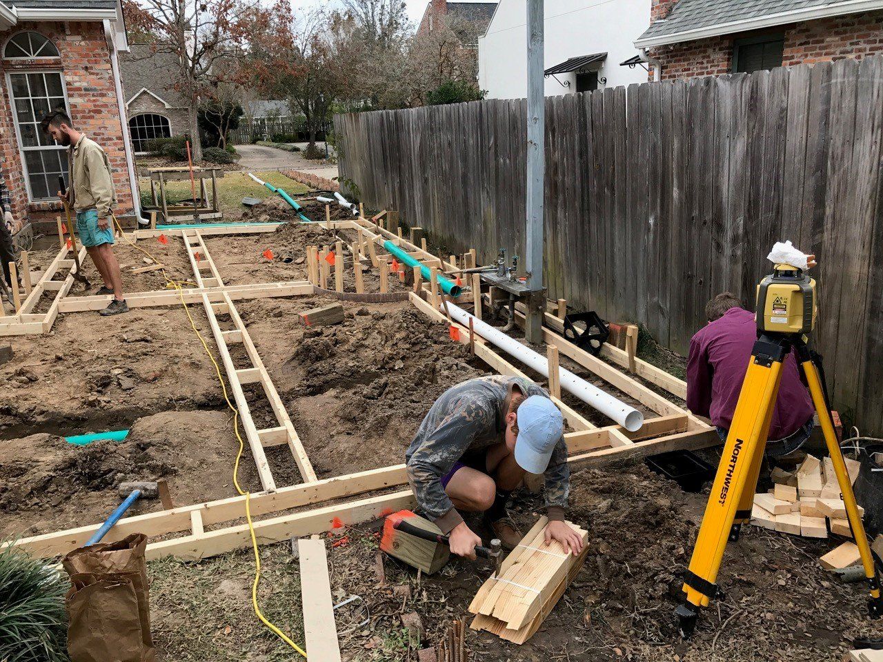 Construction site: workers building a wooden structure outdoors. A level is on a tripod; pipes and dirt are present.