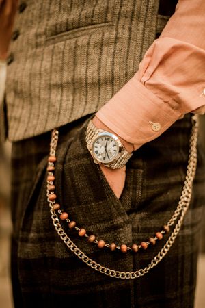 Close-up of a person's hand in a pocket, wearing a watch. A vest, pink shirt, and chain are also visible.