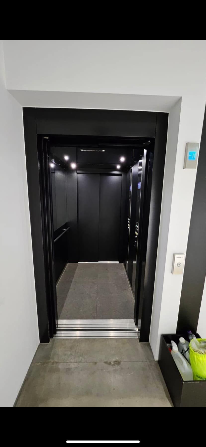 Elevator with black interior and trim. Grey floor. White walls. Bright ceiling lights.