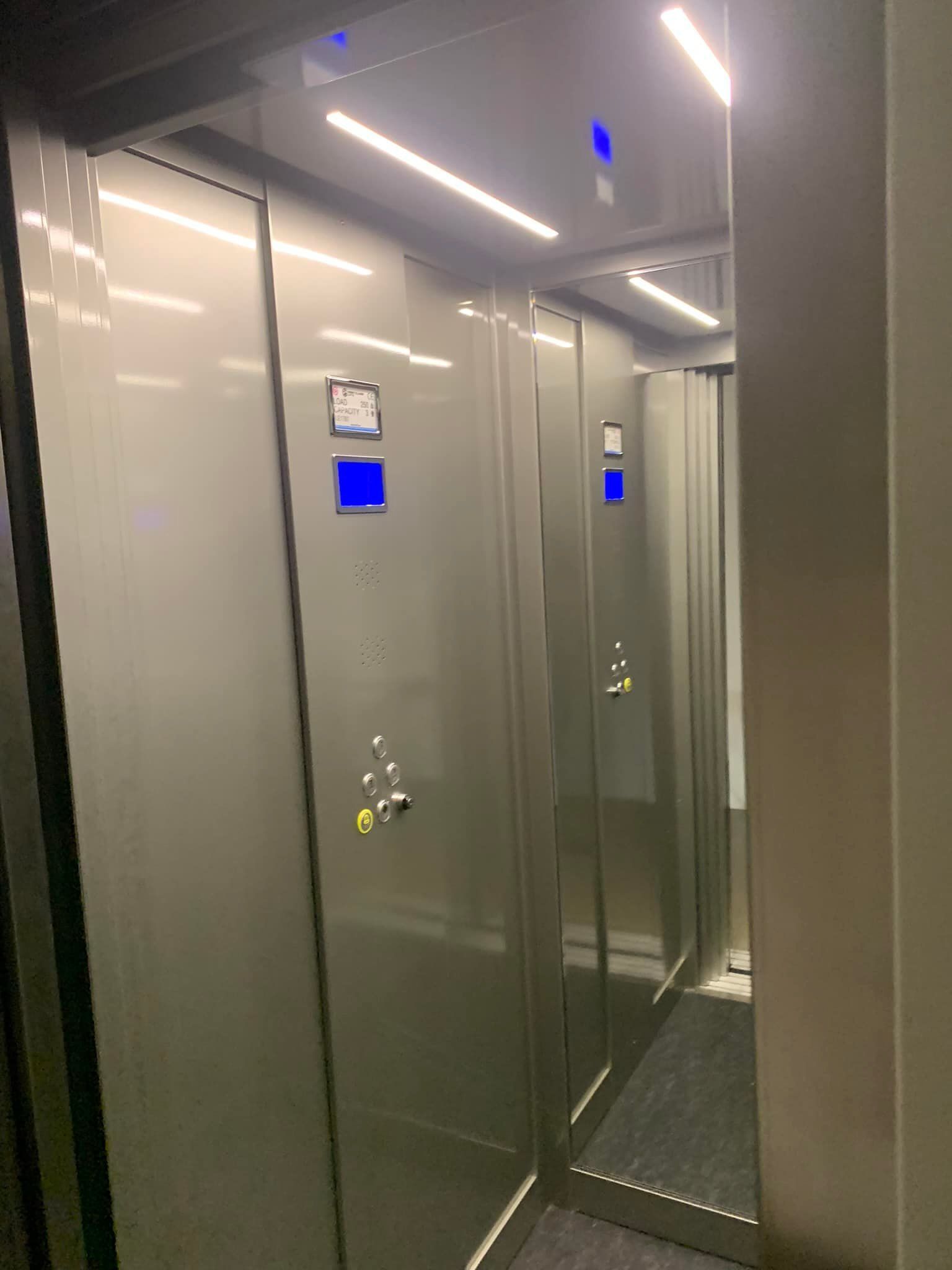 Elevator interior with metallic gray doors, blue indicator lights, and overhead lighting.