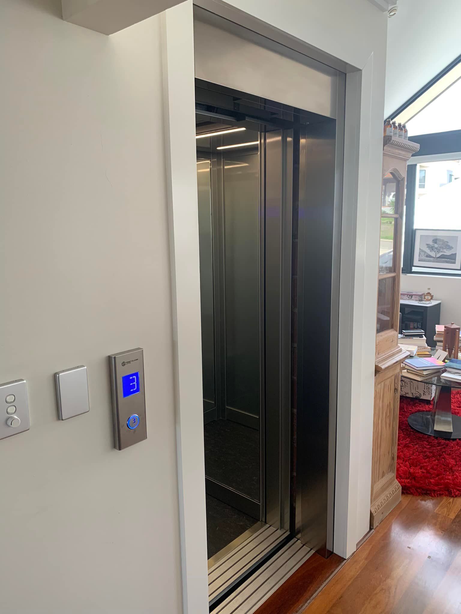 Elevator with stainless steel door and control panel. White walls, wooden floor, and red rug visible.