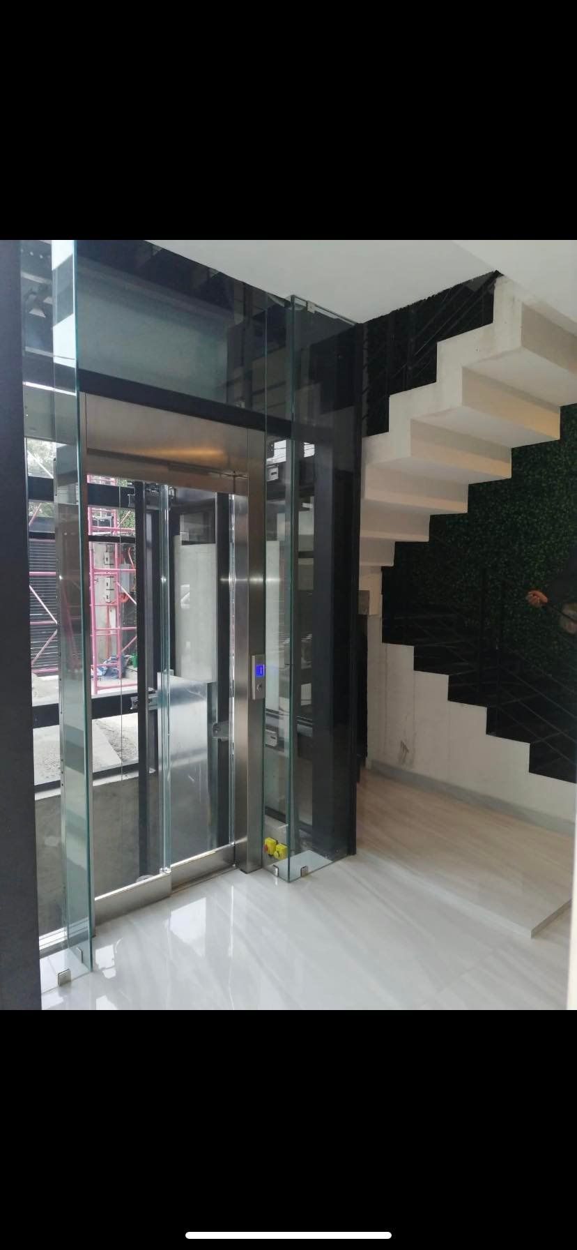 Modern glass elevator next to a staircase. Light-colored tiles on the floor. Black walls.