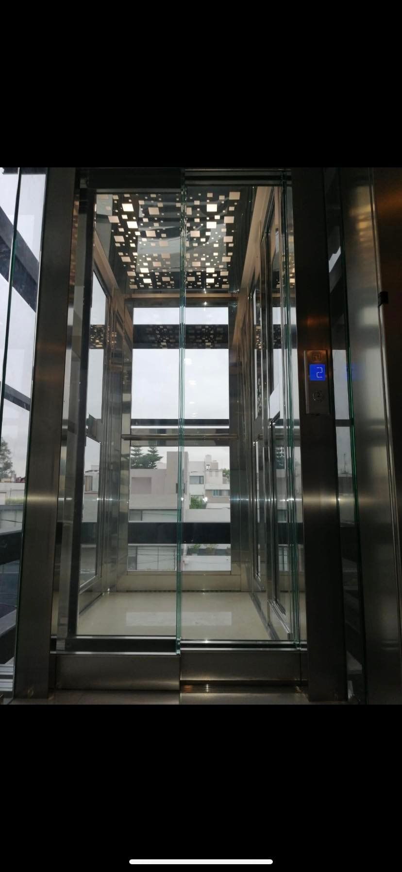 Interior view of a modern glass elevator. Shows shiny metal framing with glass panels, reflecting the outside view.