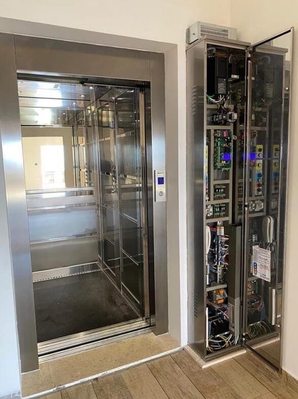 Elevator with open door, stainless steel interior. Electrical panel cabinet is open, revealing wiring and components.