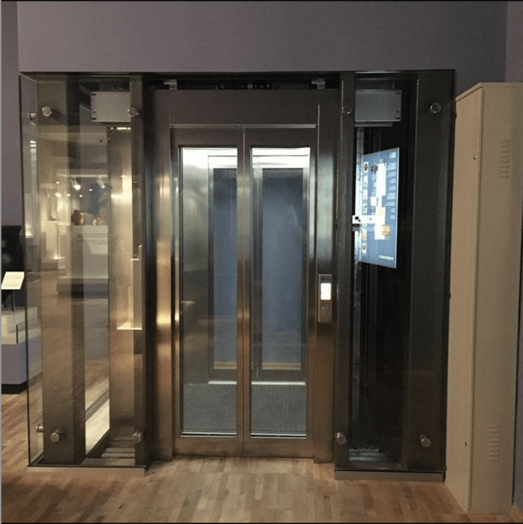 Elevator with glass and stainless steel exterior, interior access panel, and a small screen.