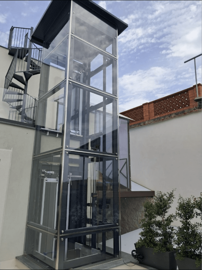 Exterior elevator with glass walls and metal frame next to spiral staircase and building.