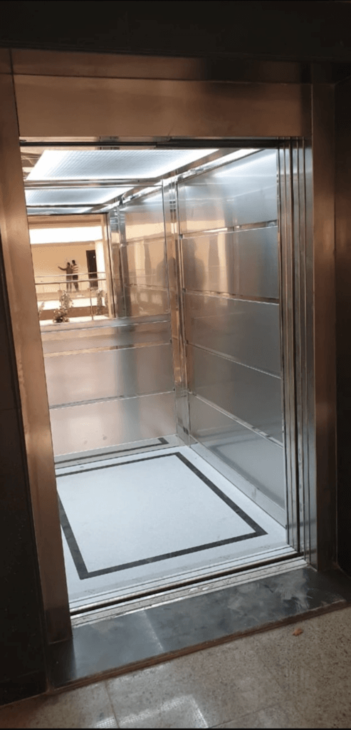 An empty, modern elevator with stainless steel walls and a white floor with a black border.