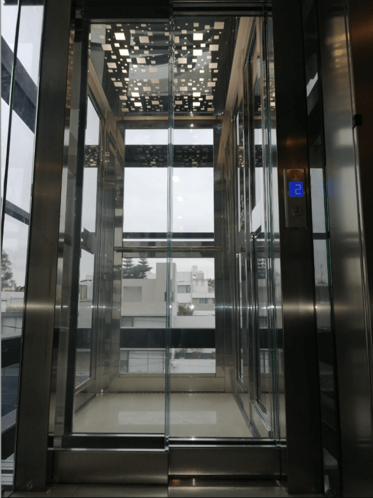 Glass elevator with open door, showing exterior view. Interior has stainless steel frame, patterned ceiling.