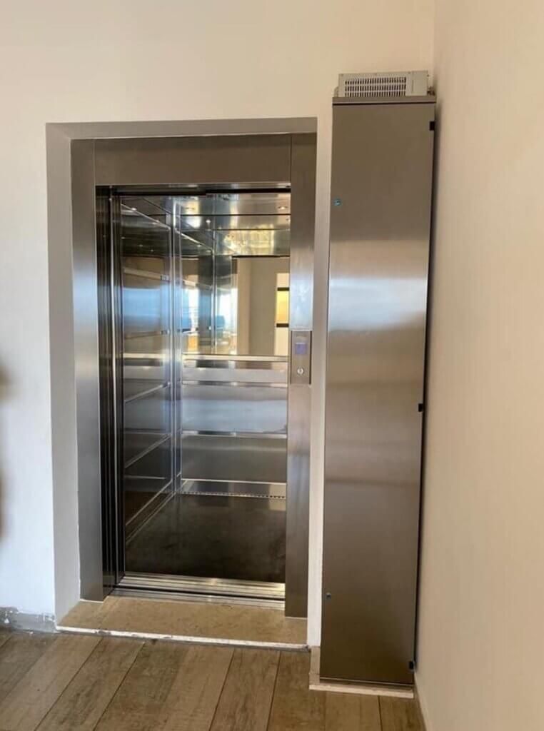 Elevator with stainless steel door and control panel. Doorway framed in gray. Adjacent wall with a panel.