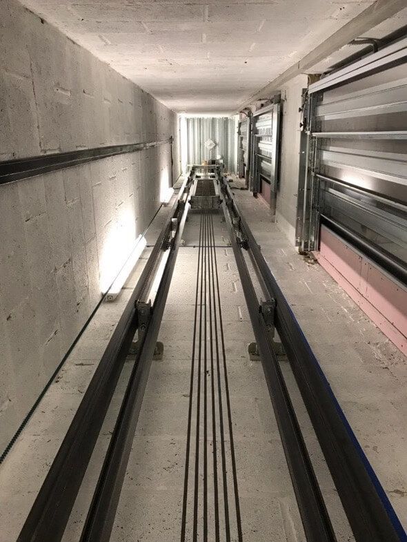 Elevator shaft with guide rails, cables, and open doors at both ends, concrete walls, interior view.