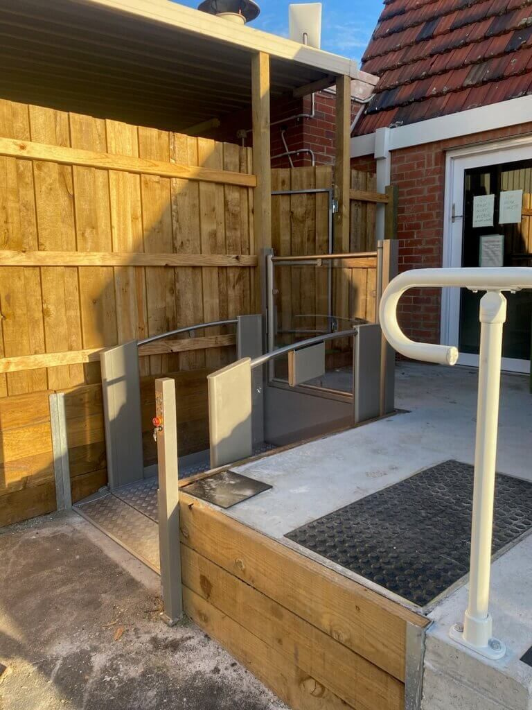 Exterior view of a ramp and lift with handrails, leading to a building entrance. Wooden fence and brick wall visible.
