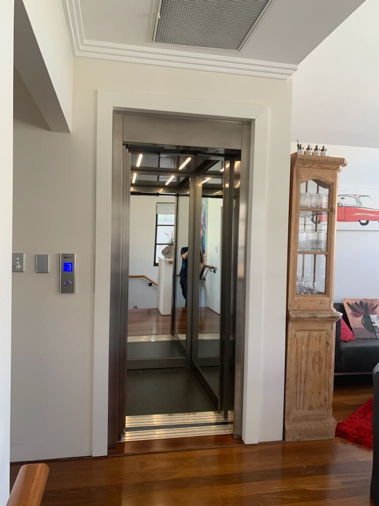 Elevator in a home. Open stainless steel doors, interior mirrors, and a person reflected inside. Wooden floors and walls.