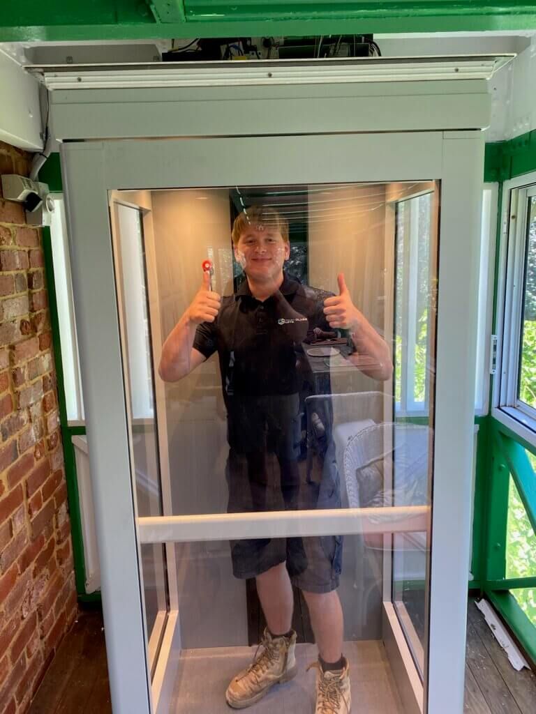 Man giving thumbs up inside a glass elevator with a white frame. Green and brick surroundings.