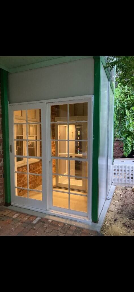 White-framed glass doors of a small building with green trim and a red brick floor, surrounded by greenery.